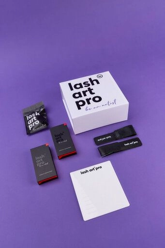 A collection of eyelash extension products on a purple background. Items include a white box labeled lash art pro, black product boxes, adhesive containers, black tweezers in sleeves, and a notepad.